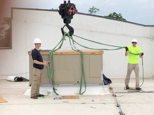 Two MLD technicians in hard hats guiding a crane as it lowers a rooftop commercial HVAC unit into place.
